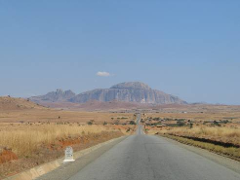 A long, paved road stretches towards a distant mountain range under a clear blue sky, with dry grassland flanking both sides.