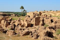 The image shows ancient ruins with crumbling brick structures in the foreground, backed by a restored brick wall with crenellations and palm trees against a clear blue sky.