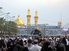 A large crowd gathers in front of a grand, ornate mosque with golden domes and minarets under a cloudy sky.