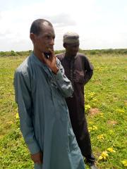 Two men stand in a field of yellow flowers under a cloudy sky, with the man in front wearing a blue robe and the other in a dark robe with a hat.