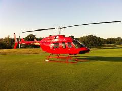 A red helicopter sits on a green grass field with trees and a clear sky in the background.