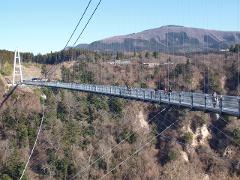 A long suspension bridge spans a valley, with people walking on it against a backdrop of forested mountains and a clear blue sky.