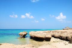 A serene coastal landscape with turquoise waters, unique rock formations, and a small thatched hut under a bright blue sky with scattered clouds.
