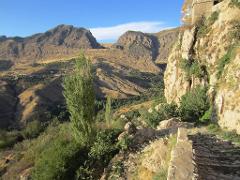 Une vue panoramique d&#39;un terrain montagneux avec un chemin de pierre le long d&#39;une falaise, avec des arbres et un bâtiment en haut à droite, le tout sous un ciel bleu.