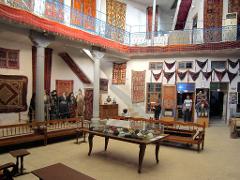 The image shows an interior space filled with carpets, traditional clothing on mannequins, and furniture, reminiscent of a museum or historical exhibition.