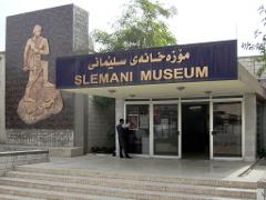 The Slemani Museum entrance is captured in a daytime shot, featuring a man placing a poster on the glass doors under a sign with Arabic and English text, flanked by a stone carving.