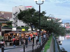 A lively riverside scene unfolds with a bustling market along a canal, lined with food stalls and people strolling on the pavement, set against a backdrop of buildings and a sky hinting at dusk.