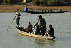 A group of four men are crossing a river in a small boat, along with a motorbike, in front of a larger boat and a flag on the shore.