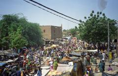 A bustling outdoor market scene in an African town with a large crowd of people, various stalls, trees, and a building with small windows.