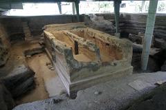 An archaeological site featuring exposed stratified layers of earth and the remains of a building with walls, showcasing an excavation area under protective structures.