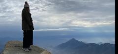 A person stands on a rocky peak overlooking a misty mountain range beneath a cloudy sky in the Asir region.