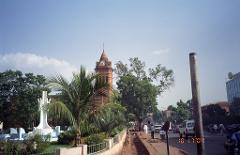 A vibrant street scene featuring a clock tower partially obscured by lush greenery, with a streetscape bustling with pedestrians, vehicles, and a clear blue sky above.