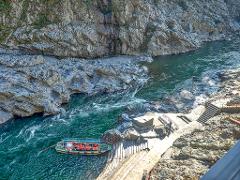 Una toma aérea captura una garganta de un río con un bote lleno de pasajeros con chalecos salvavidas naranjas, atracado cerca de unas escaleras que conducen a una pasarela pavimentada junto a acantilados rocosos.