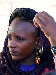 A woman with striking tribal tattoos and elaborate earrings gazes thoughtfully to the left while adjusting her headscarf and displaying beaded bracelets.