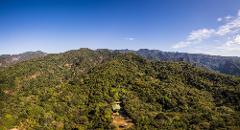 An aerial shot shows a lush, green mountain range under a bright blue sky, with a small building nestled among the trees on the hillside.
