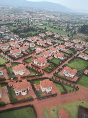 An aerial shot shows a wealthy residential neighborhood with large houses, red tile roofs, manicured lawns, and trimmed hedges, set against a backdrop of distant city and mountains.