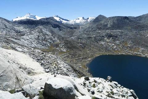 Sierra Nevada Cultural Immersion Experience (4 Days / 3 Nights) A panoramic view of a vast, rugged mountainous landscape under a clear blue sky, featuring snow-capped peaks in the distance, rocky terrain, and a dark, still lake in the foreground.