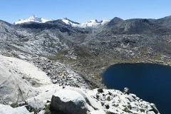 Sierra Nevada Cultural Immersion Experience (4 Days / 3 Nights) A panoramic view of a vast, rugged mountainous landscape under a clear blue sky, featuring snow-capped peaks in the distance, rocky terrain, and a dark, still lake in the foreground.