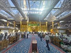 The Imam Ali Mosque shines under a glass ceiling, its golden walls and pillars reflecting light amidst a crowd of people on a blue carpet.