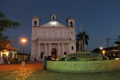 At night, children peer into a tiled fountain near a large white church in a plaza illuminated by streetlights.
