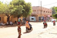 A street scene shows a woman carrying goods on her head, motorcycles driving by, and men sitting and conversing under a tree in a sunny, dusty African town.