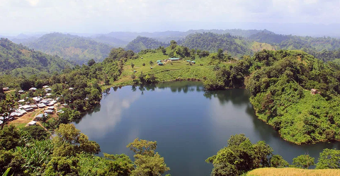 A scenic aerial view of a serene lake nestled amidst lush green hills and vegetation, with small settlements dotting the landscape.