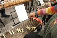 A person in a colorful, patterned sleeve plays a koto, a traditional Japanese stringed instrument, with finger picks, while another koto sits in the background.