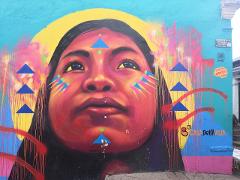 A colorful mural depicts a woman with vibrant face paint and geometric shapes, set against a turquoise background with a yellow halo, graffiti tags, and the words "Casa Bella Vista."