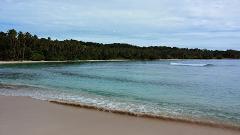 The image shows a tranquil tropical beach with clear turquoise water, a sandy shore, and lush green vegetation along the coastline under a partly cloudy sky.