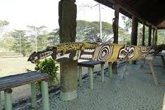 A decorated wooden sculpture, possibly a crocodile, rests on stone columns under a covered walkway in Port Moresby, with greenery and open space visible in the background.