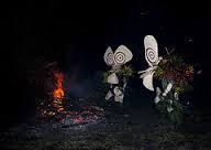Certainly! Here's a description of the image:Two figures in traditional garb with large, white headpieces stand near a burning pyre, likely part of a cultural performance or memorial ceremony in Rabaul.