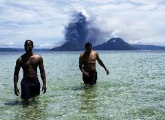 Two shirtless men wade through crystal-clear water, with an erupting volcano dramatically rising in the background, under a partly cloudy sky.
