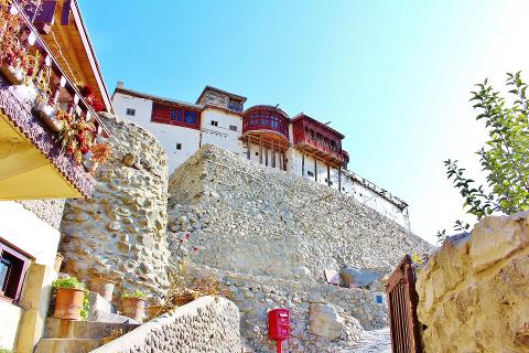 A historic white building with red accents sits atop a stone wall, with a red mailbox and greenery in the foreground under a bright blue sky.