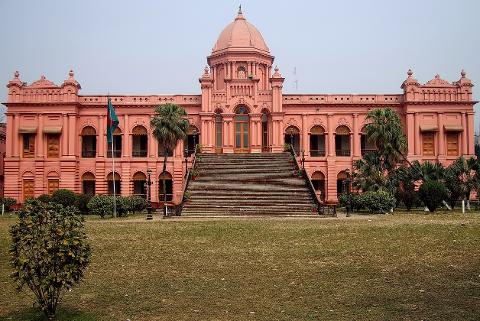 Bangladesh Visa on Arrival for Chinese Citizens: Complete Guide A symmetrical pink building with a dome sits behind a large lawn and steps, featuring a flag to the left and various trees and bushes.