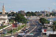 An aerial view of a busy road in Libreville, Gabon, shows traffic, buildings, and lush greenery under a clear sky.