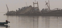 A dilapidated shipwreck sits in shallow water, with a small boat in the foreground and a hazy sky above.