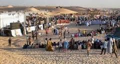 Here is a description of the image in one sentence:A bustling crowd gathers on sandy ground with makeshift stages and structures, surrounded by desert dunes, likely during the Agadez African Music Festival.