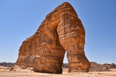 A massive, arch-shaped rock formation dominates a desert landscape under a clear blue sky.
