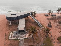 An aerial view shows an abandoned beach resort with a unique, circular structure connected to a walkway, palm trees, and people on the sandy ground.