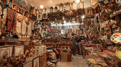 A cluttered shop displays a variety of handcrafted items, from framed art and lanterns to metal shields and trinkets, with two customers browsing.