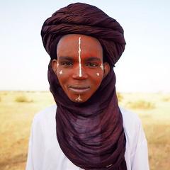 A Tuareg man with traditional face paint and indigo turban stands in a desert landscape, possibly during the Gerewol festival or Cure Salee.