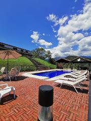 A serene outdoor pool area with lounge chairs and umbrellas, set against a backdrop of lush greenery, blue sky, and puffy white clouds.