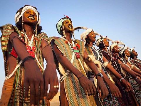 A group of Wodaabe men in traditional attire and face paint, participating in the Gerewol Festival, stand in a line with arms extended, under a clear sky.