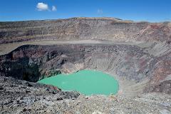 A wide shot of a large volcano crater with a green lake inside, surrounded by rocky cliffs under a clear blue sky.