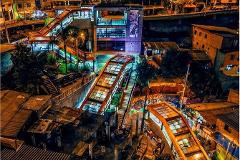 Medellín Cultural & Nightlife Experience (3 Days / 2 Nights) An aerial nighttime view shows illuminated outdoor escalators winding through a vibrant urban neighborhood with colorful murals and houses.