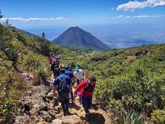 A group of people hikes up a rocky trail on a sunny day, with a large mountain and scenic view in the background.