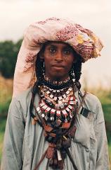 A person with tribal markings, wearing a colorful headwrap and elaborate necklaces, stands out against a natural background.