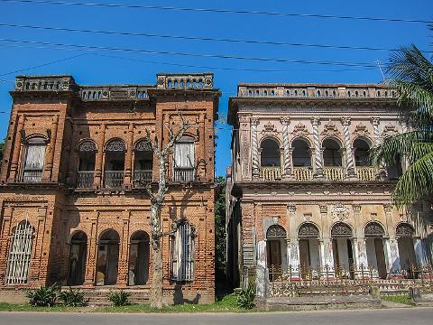 Two old, ornate brick buildings stand side-by-side under a bright blue sky, showing their age and architectural details.