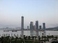 A city skyline with several skyscrapers, some under construction, alongside a body of water with ships and a waterfront park.