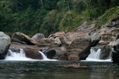 Katanzama Indigenous Community Hiking Tour with Poza Sagrada Visit A river flows over rocks, forming small waterfalls, with lush greenery in the background.